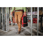 Worker carrying the M12 PACKOUT Flood Light with USB Charging in a construction site, with additional tools and equipment in the background.