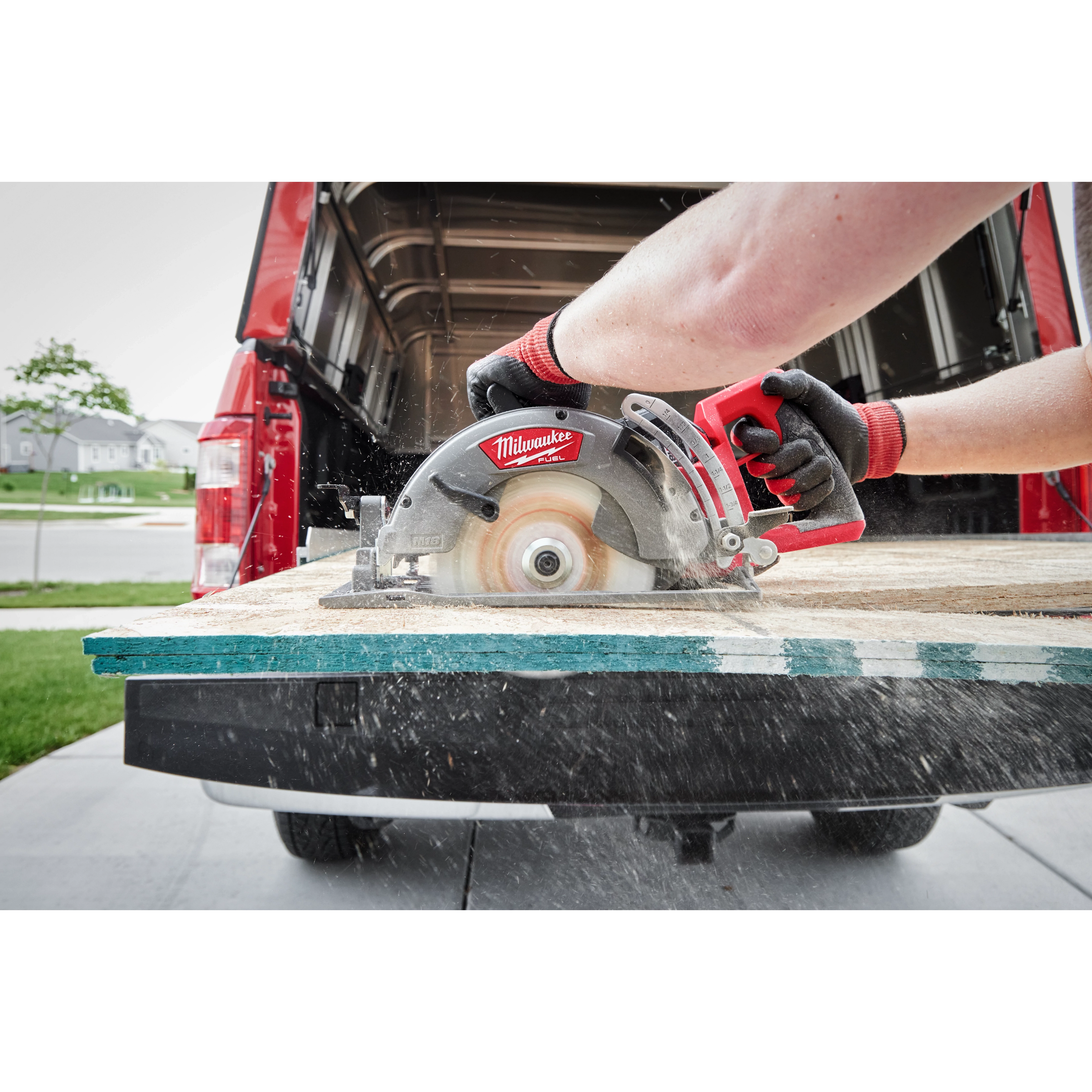 A person uses the M18 FUEL™ Rear Handle Circular Saw to cut plywood at the edge of a pickup truck bed. The saw is in action, producing wood shavings, with a suburban neighborhood visible in the background.