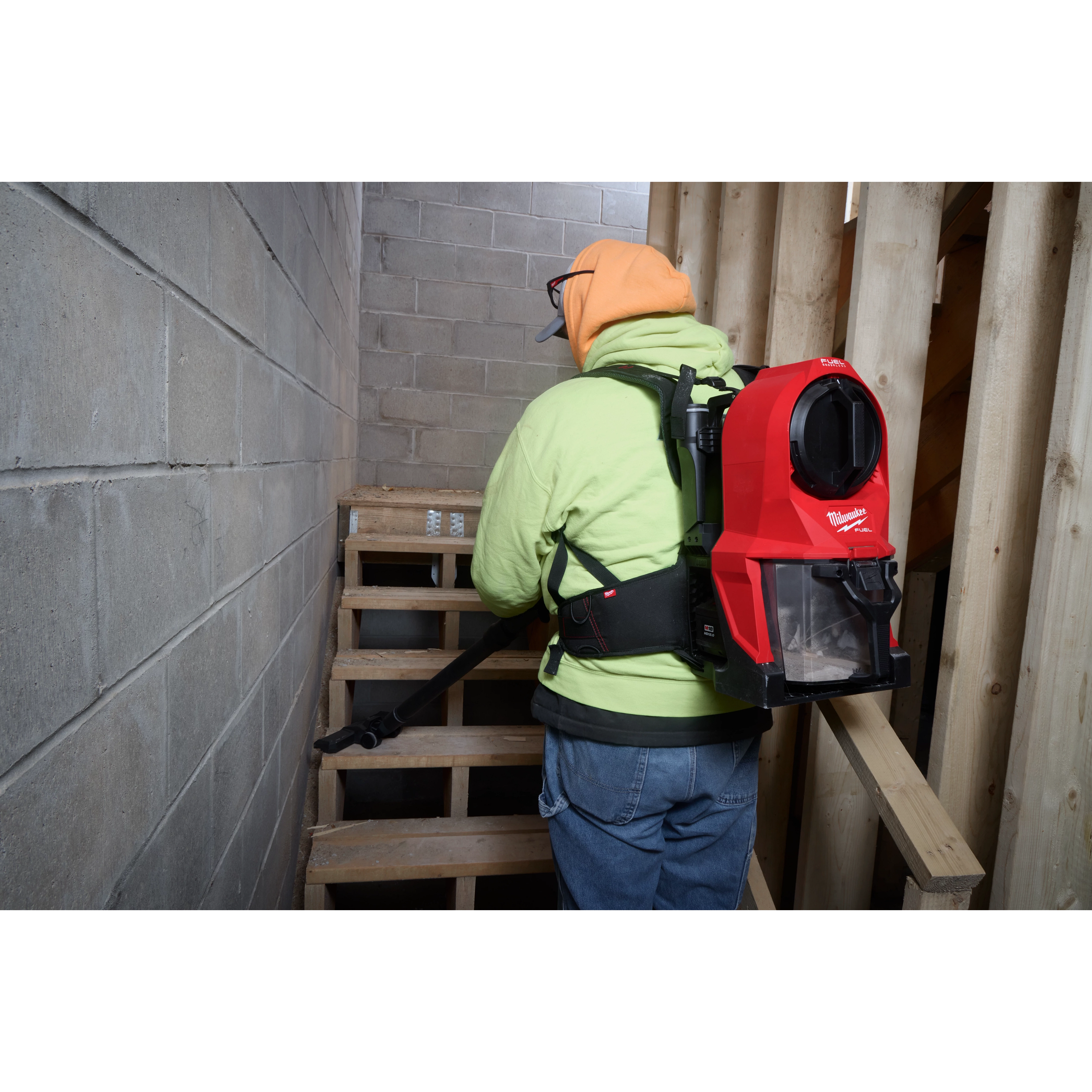 A person wearing a neon green hoodie and orange cap is using the M18 FUEL™ 3-in-1 Backpack Vacuum. They are cleaning a wooden staircase in a construction setting, with the red and black vacuum secured on their back.