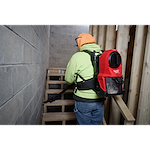 A person wearing a neon green hoodie and orange cap is using the M18 FUEL™ 3-in-1 Backpack Vacuum. They are cleaning a wooden staircase in a construction setting, with the red and black vacuum secured on their back.