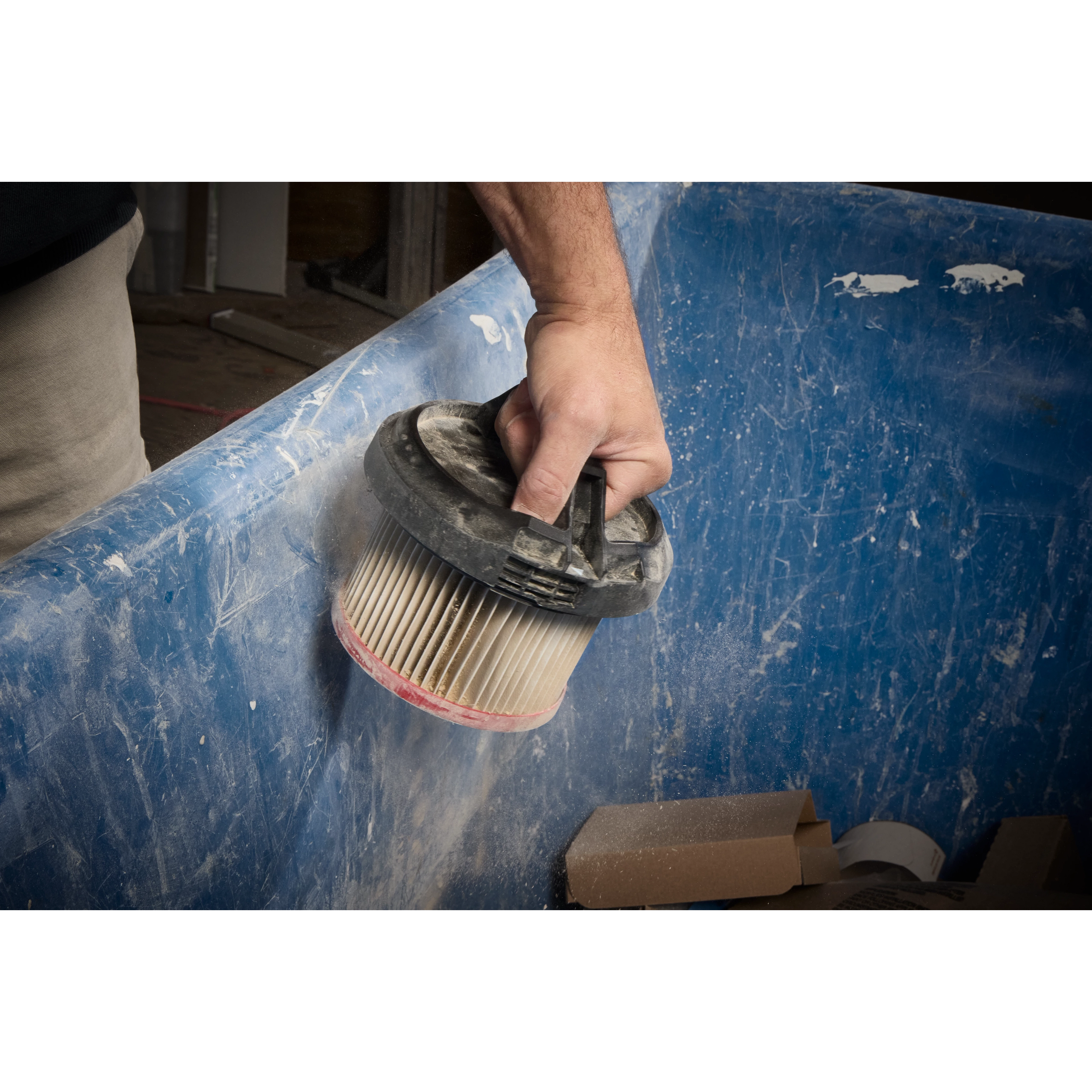 A person holds a dust-covered filter from the M18 FUEL™ 3-in-1 Backpack Vacuum over a blue container, likely for cleaning or replacement.