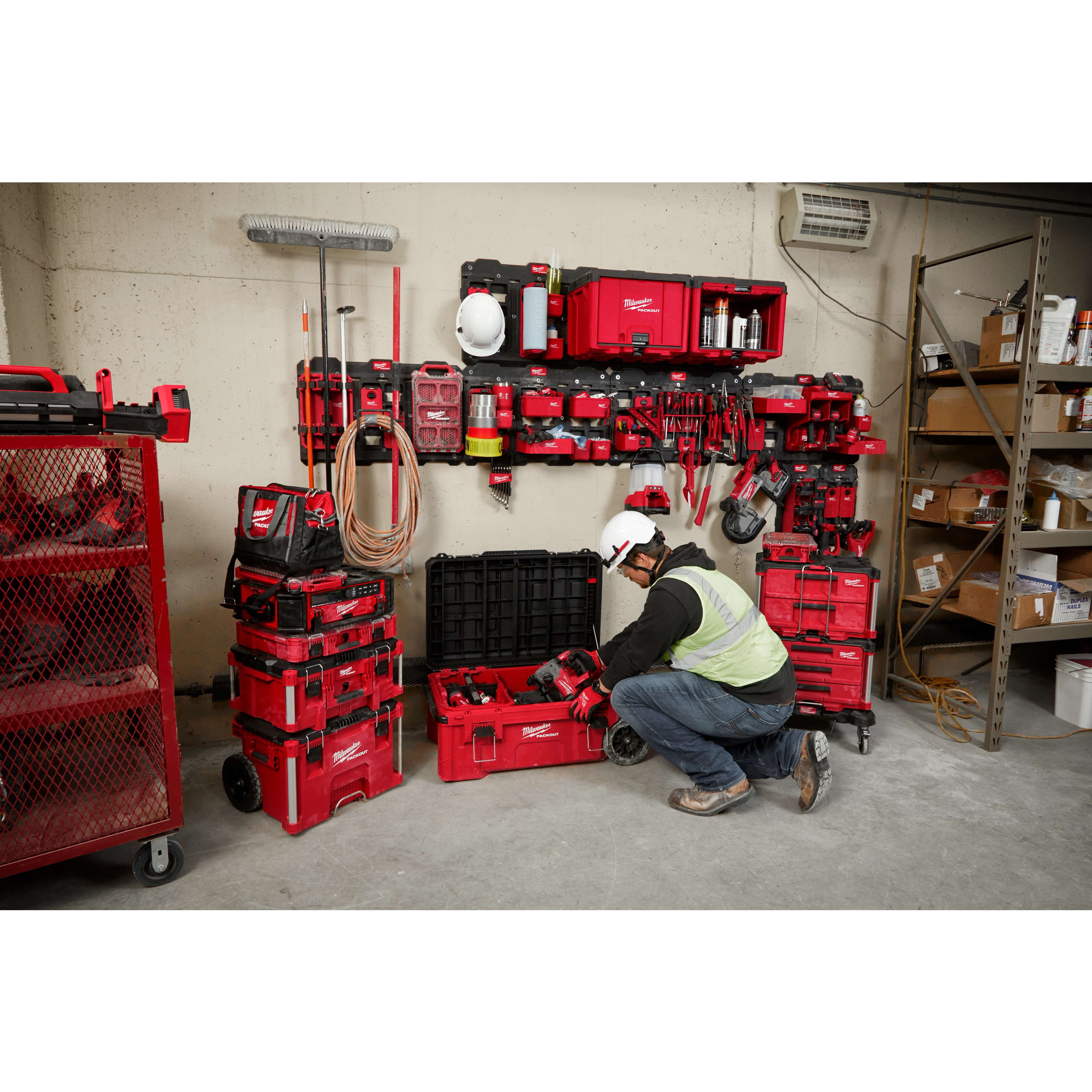 A worker in a hard hat and safety vest organizes tools with the PACKOUT™ Shop Storage Breadth of Line Image system, comprising red storage boxes and wall-mounted units filled with various tools and accessories. The system is designed for efficient organization on a shop floor.
