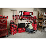 A worker in a hard hat and safety vest organizes tools with the PACKOUT™ Shop Storage Breadth of Line Image system, comprising red storage boxes and wall-mounted units filled with various tools and accessories. The system is designed for efficient organization on a shop floor.