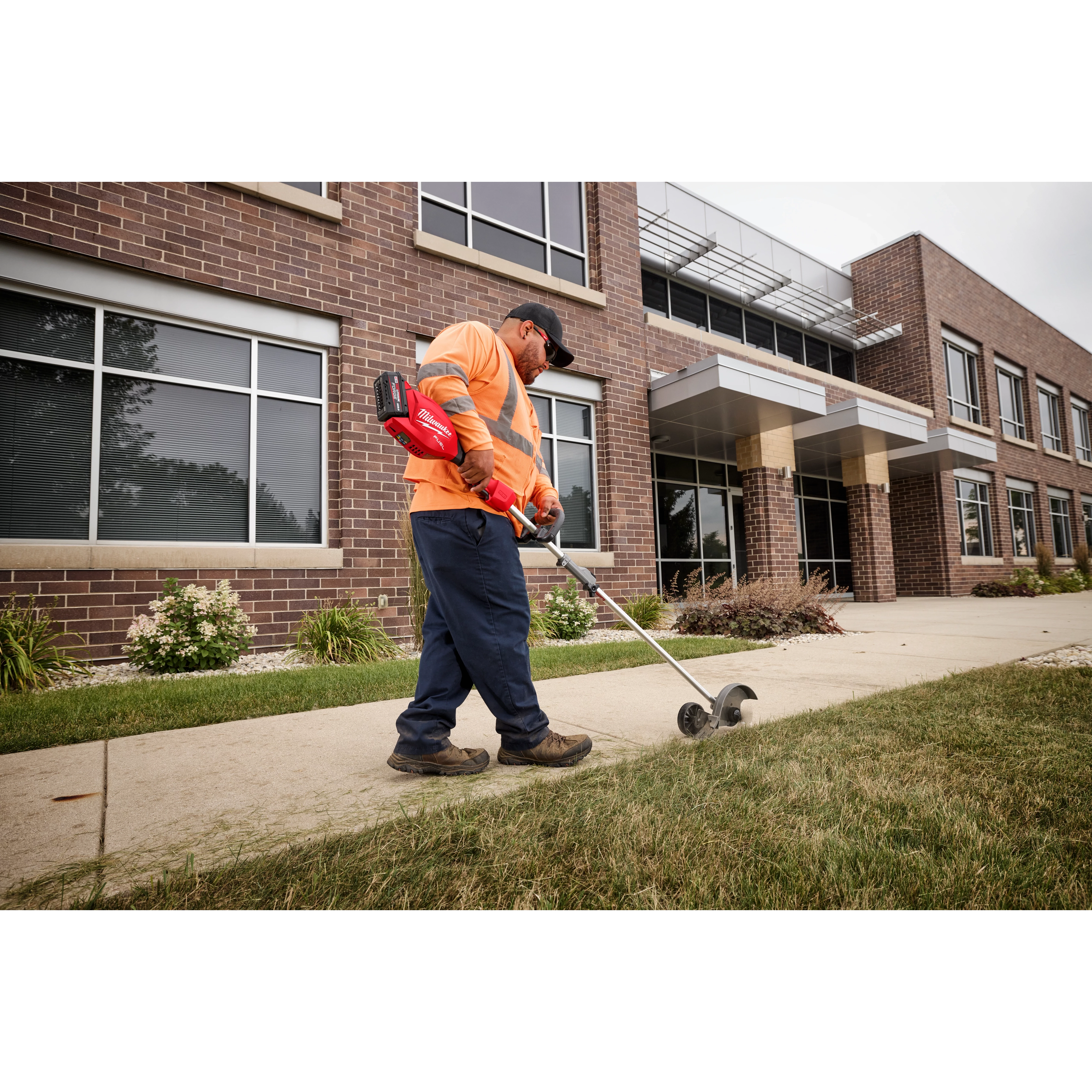 Worker with trimmer edges lawn near brick building with large windows and canopy, wearing orange shirt and blue pants.