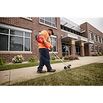 Worker with trimmer edges lawn near brick building with large windows and canopy, wearing orange shirt and blue pants.