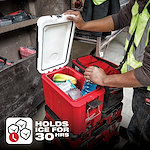 The 48-22-8460%20App%20Shot is a red, insulated cooler box with an open lid, revealing ice, drinks, and snacks inside. A person wearing a reflective vest is accessing the cooler. A label indicates it can hold ice for 30 hours.