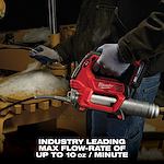 A person in gloves operates a Milwaukee power tool, specifically a cordless grease gun, in a workshop. The text at the bottom reads, "INDUSTRY LEADING MAX FLOW-RATE OF UP TO 10 oz / MINUTE." Snow is visible on the equipment being serviced.