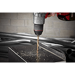 The RED HELIX™ Cobalt 3/8" Drill Bit is attached to a drill and is actively drilling into a metal surface, creating fine metal shavings. The drill is held by a hand and positioned above a square metal object, with industrial pipes in the background.