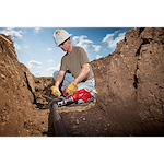 A worker uses the FORCELOGIC™ M18™ 3" Underground Cable Cutter Kit to cut a large underground cable. The tool is red and designed for heavy-duty industrial use. The worker is wearing yellow gloves, a white hard hat, and sits in a trench on a sunny day.