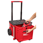 A person places a tool into a red PACKOUT™ Rolling Drawer Tool Box by Milwaukee, which has an extended handle and large wheels for easy transport.