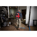 Man using the M18 FUEL™ 3-in-1 Backpack Vacuum to clean a hardwood floor in an industrial setting. The vacuum is red and black, strapped to his back, and connected through a long hose. The room contains large machinery and is partially under construction.