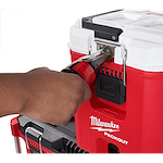 A person is using a built-in bottle opener on a red and white PACKOUT™ 16QT Compact Cooler. The cooler has the Milwaukee logo on the front and is stacked on another PACKOUT™ unit.