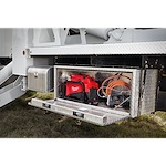 Open storage compartment on a vehicle containing an M18™ FORCE LOGIC™ 10,000 PSI Hydraulic Pump, a coiled orange hose, and additional electrical tools. The hydraulic pump is red and prominently positioned at the center of the compartment.
