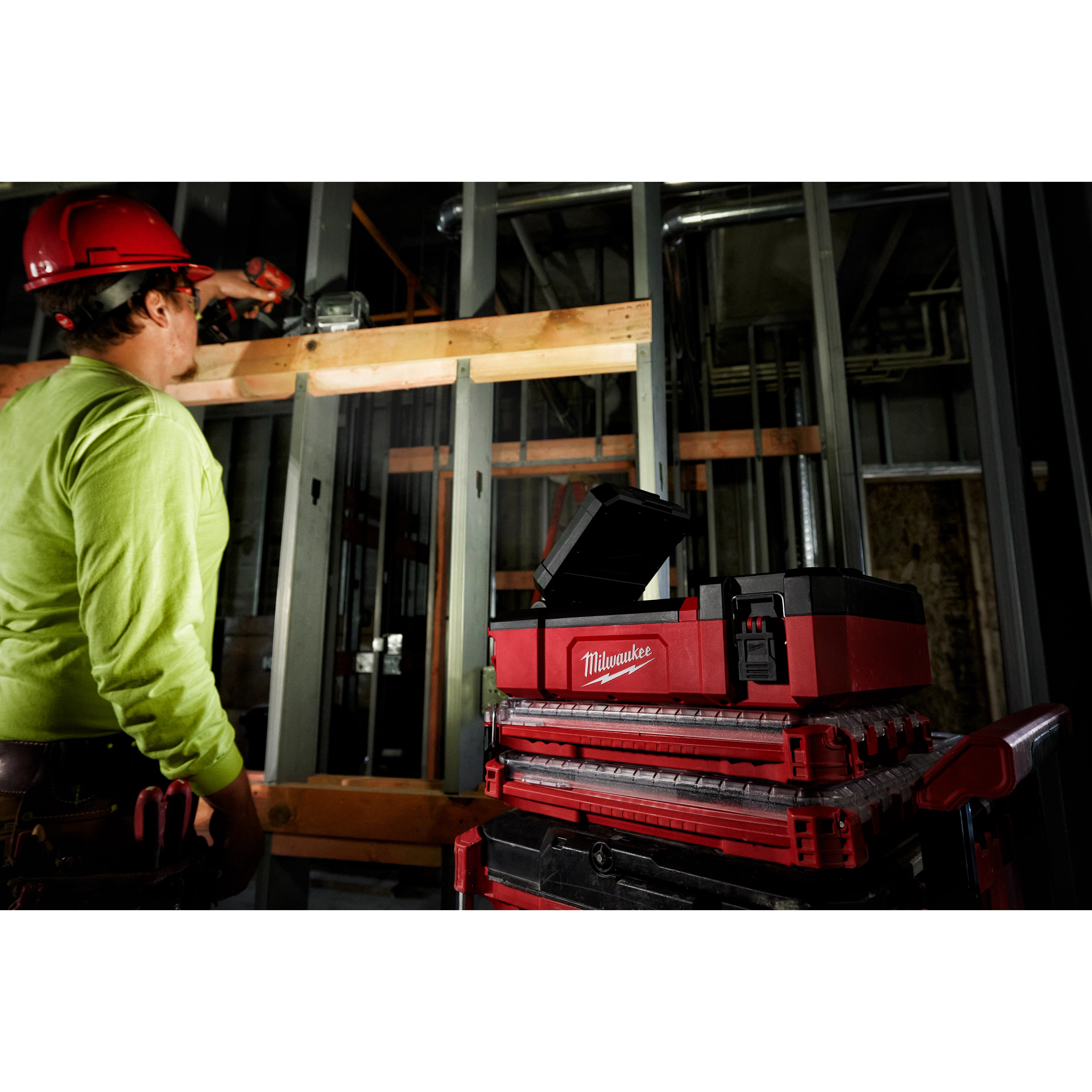 Worker using a drill at a construction site, with M12 PACKOUT Flood Light w/ USB Charging on a stack of red and black toolboxes nearby.