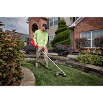 Person using a string trimmer to edge the lawn in front of a house surrounded by neatly landscaped plants.