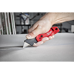 A person cutting a white material using a red FASTBACK Folding Utility Knife, with the knife held in their right hand.