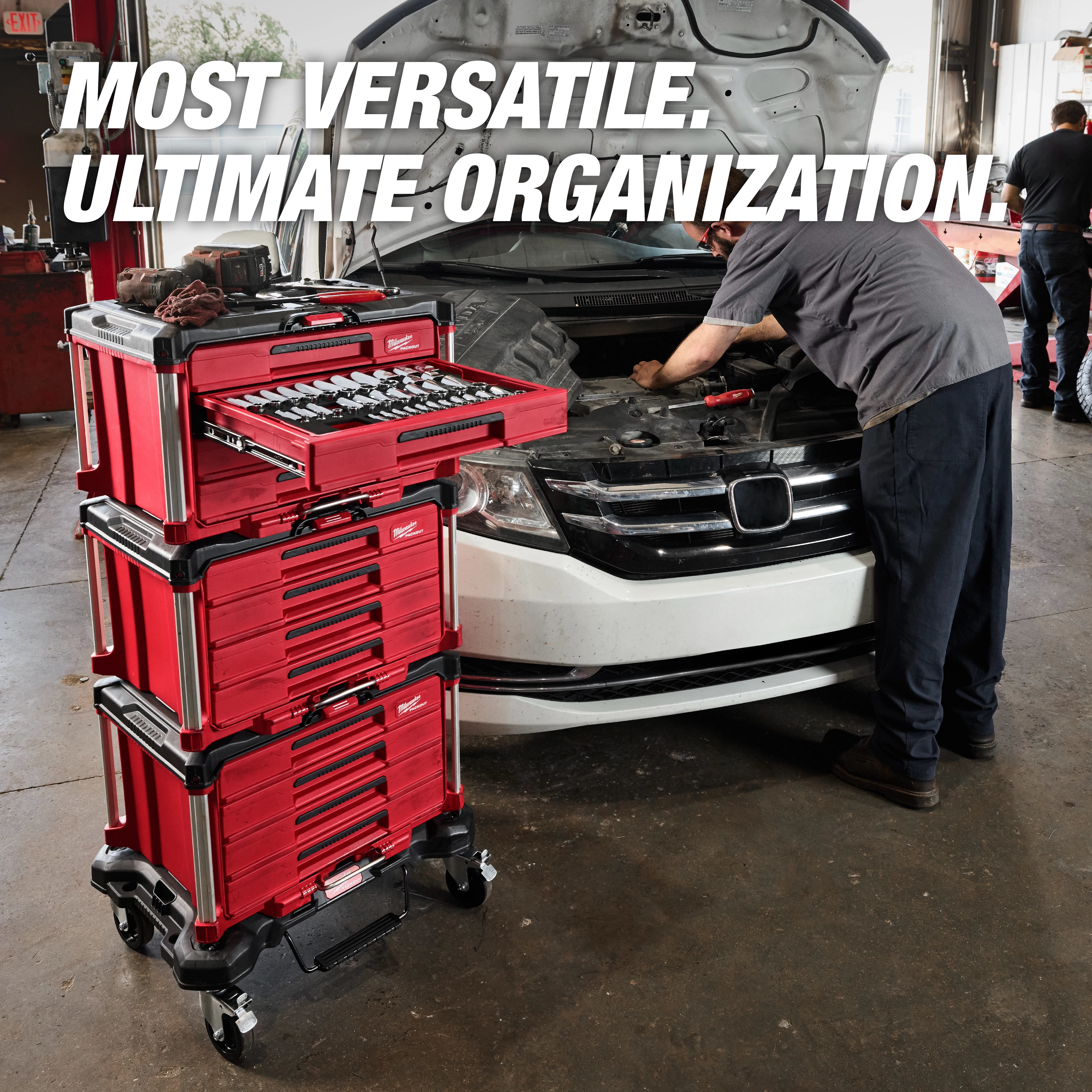 The Milwaukee Packout modular storage system is shown in a garage next to a mechanic working on a car. The red and black stacked toolboxes are labeled with the brand's logo. A drawer with tools is pulled out, and promotional text reads: "Most versatile. Ultimate organization."