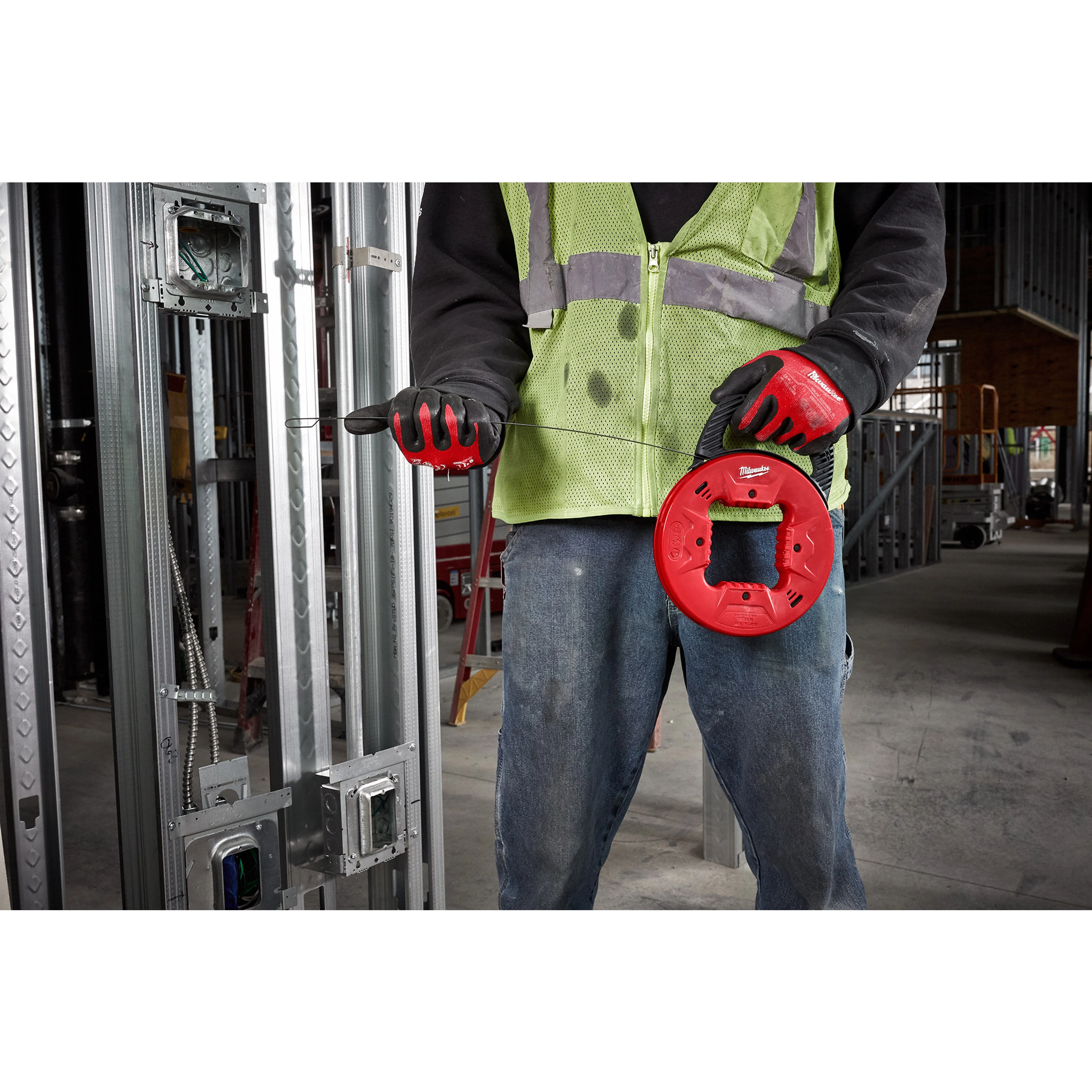 Person holding a 1/8" 50' Steel Fish Tape, wearing a hi-visibility vest and gloves, in a construction site with metal framing.