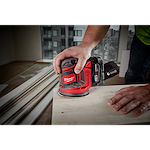 A person uses the M18 Random Orbit Sander to smooth a wooden surface. The sander has a red and black design and is cordless. The scene appears to be inside a room under construction, with visible building materials and high-rise buildings outside the window.