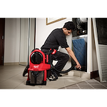 A worker uses a M18 FUEL™ 3-in-1 Backpack Vacuum to clean an HVAC system. The red vacuum is worn on the worker's back and has a black hose attached, ensuring efficient dust removal. The worker wears protective black clothing and kneels beside the HVAC unit.