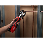 The M12 Force Logic Press Tool Kit, 2473-22, is shown being used to press a copper pipe fitting. The tool is red and black, held by a person's hand, and is pressing a connection in a wall under construction with metal studs.