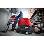 A person operates the M18 FUEL™ Rotary Hammer with Chisel Boot, M18 FUEL 1-9/16 SDS MAX Hammer Drill. The tool is red and black and is connected to a Milwaukee vacuum system for dust collection. The person wears safety gear and stands next to a construction lift.