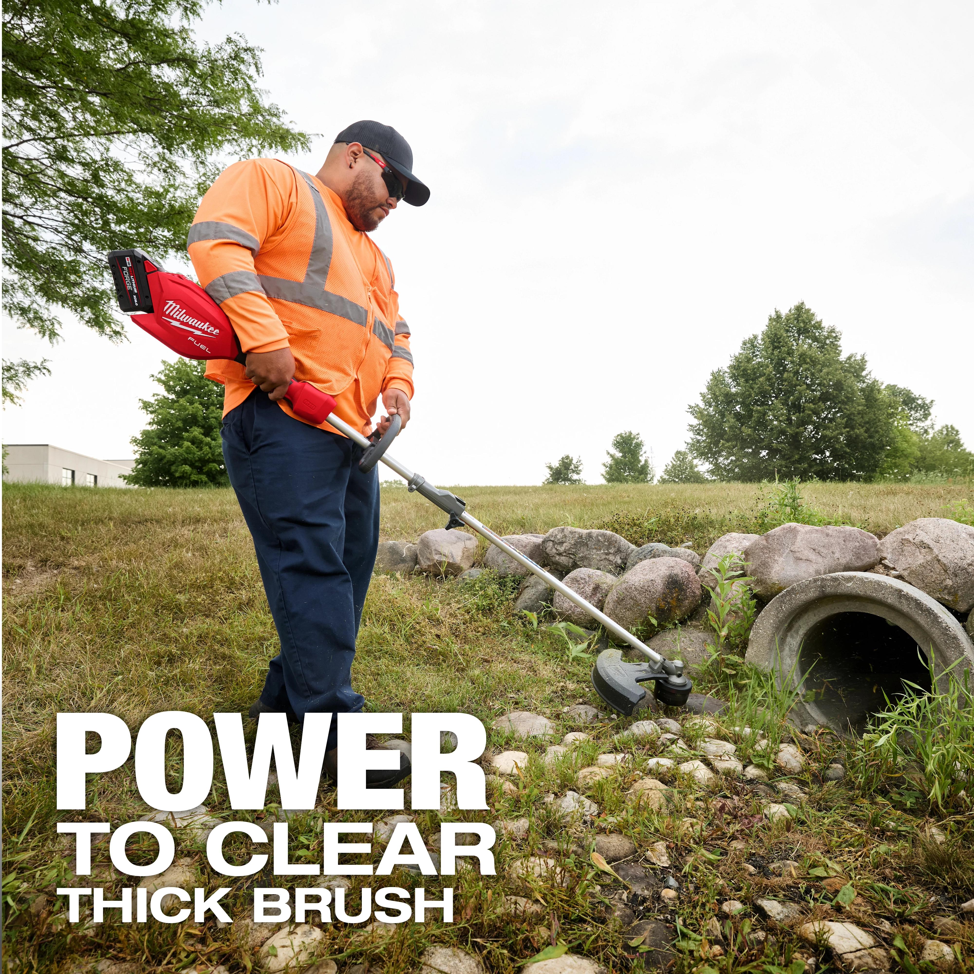 Worker using an M18 FUEL QUIK-LOK String Trimmer Attachment to cut grass near rocks and a drainage pipe.