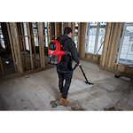 A person uses an M18 FUEL™ 3-in-1 Backpack Vacuum to clean a construction site. The individual wears the red backpack vacuum and operates the hose attachment to clean the floor in a partially framed building.