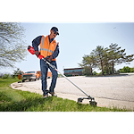 A person wearing an orange vest and using an M18 FUEL™ Power Head w/ QUIK-LOK™ trimmer adjusts the grass edge on a lawn near a parking lot with trees and vehicles in the background. The person stands on a grassy area edging along the pavement.