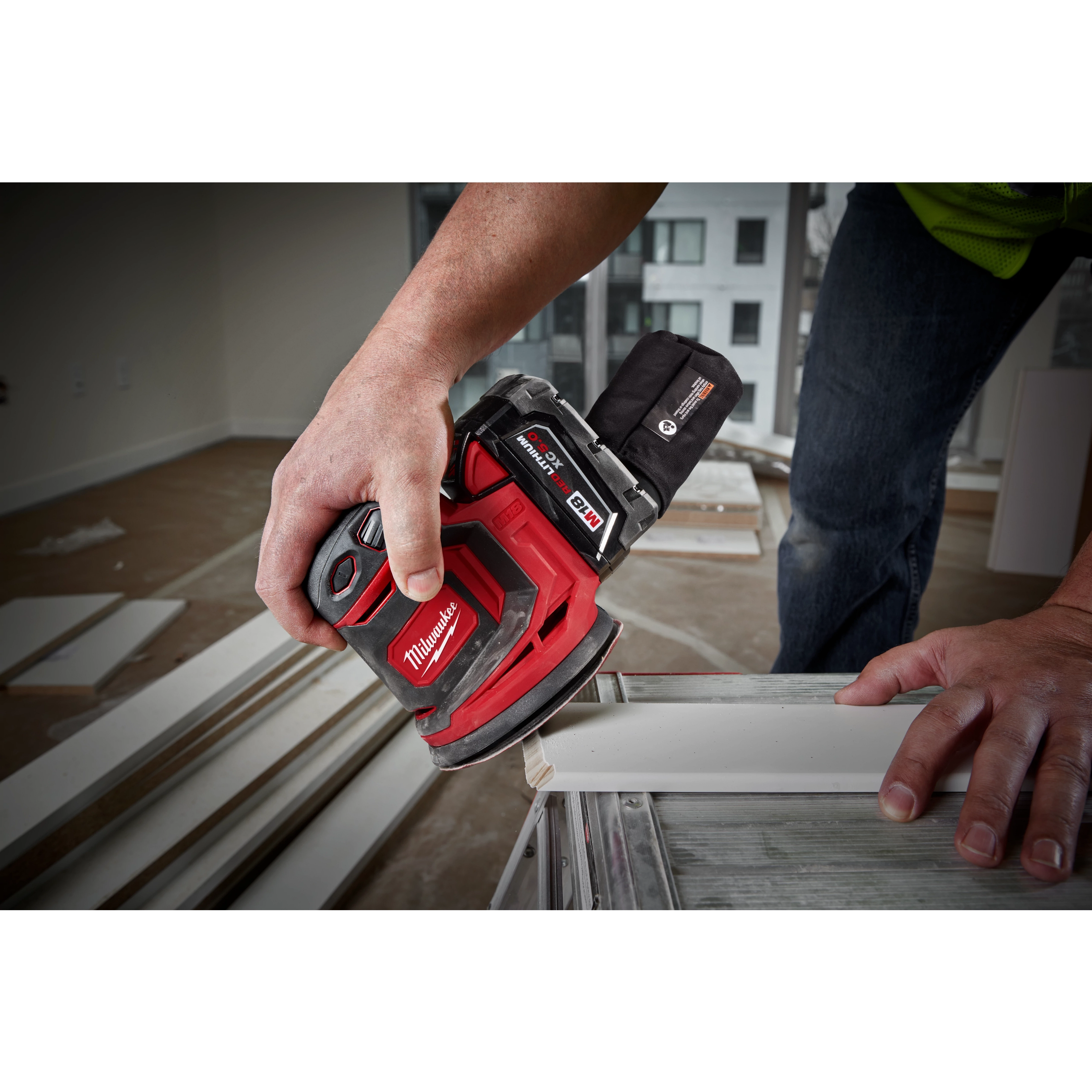 A person is using the M18 Random Orbit Sander on a piece of wood indoors. The sander is red with black accents and has a dust collection bag attached. The user is holding it firmly as they smooth the surface of the board. Additional wood pieces are visible in the background.