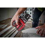 A person is using the M18 Random Orbit Sander on a piece of wood indoors. The sander is red with black accents and has a dust collection bag attached. The user is holding it firmly as they smooth the surface of the board. Additional wood pieces are visible in the background.