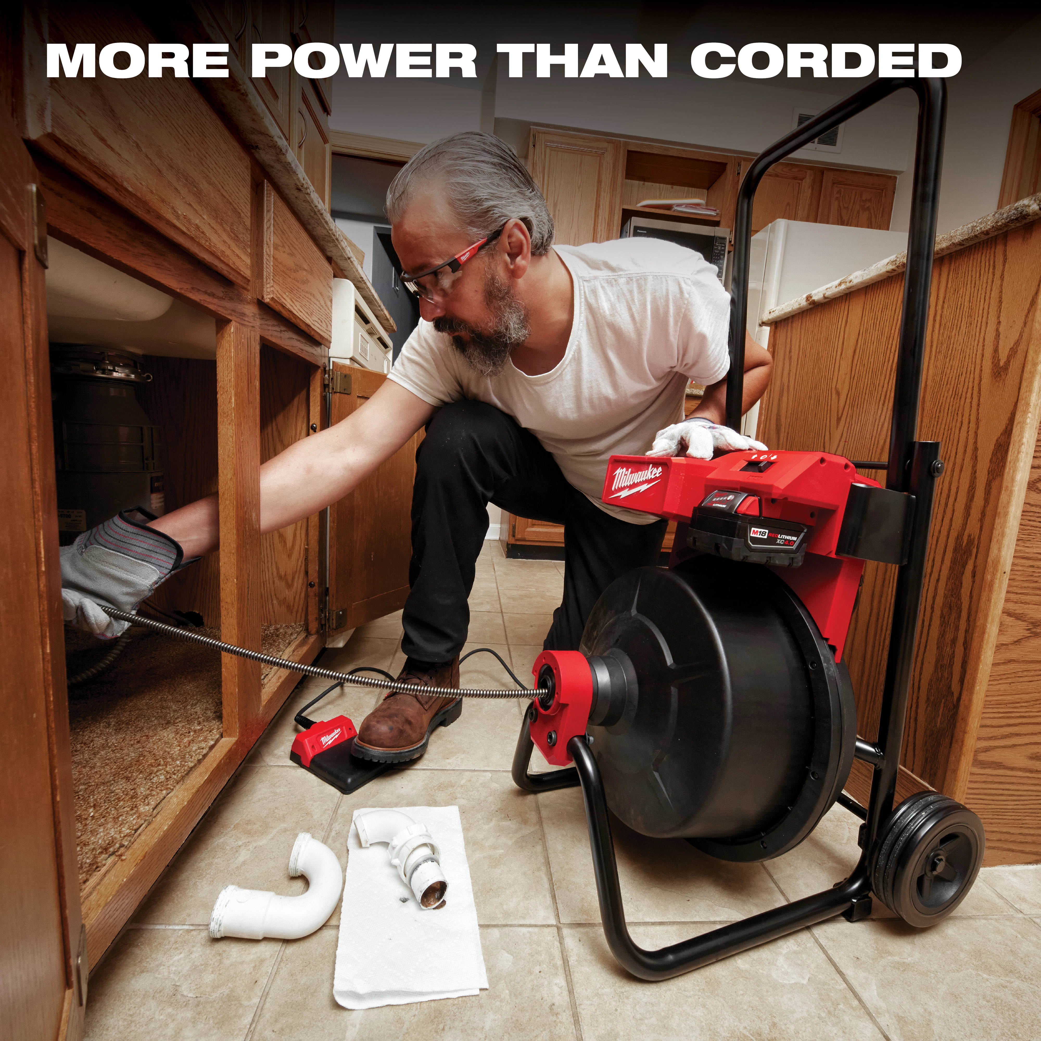 The Milwaukee MX FUEL Drum Machine is shown in a kitchen, being used to unclog pipes under a sink. A person operates the device, which features a large drum and handles, with plumbing components scattered nearby on the tiled floor. Text above reads, "More power than corded."
