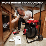 The Milwaukee MX FUEL Drum Machine is shown in a kitchen, being used to unclog pipes under a sink. A person operates the device, which features a large drum and handles, with plumbing components scattered nearby on the tiled floor. Text above reads, "More power than corded."