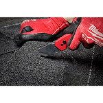 A person wearing red gloves is cutting through shingles with the FASTBACK Folding Utility Knife on a dark, rough surface.